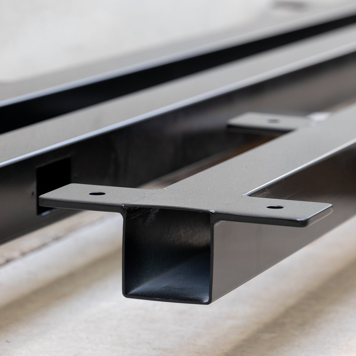 Close-up of a black metal mounting bracket or channel rail with a square tube cross-section and flat mounting flanges, photographed on a light neutral floor against a light wall.