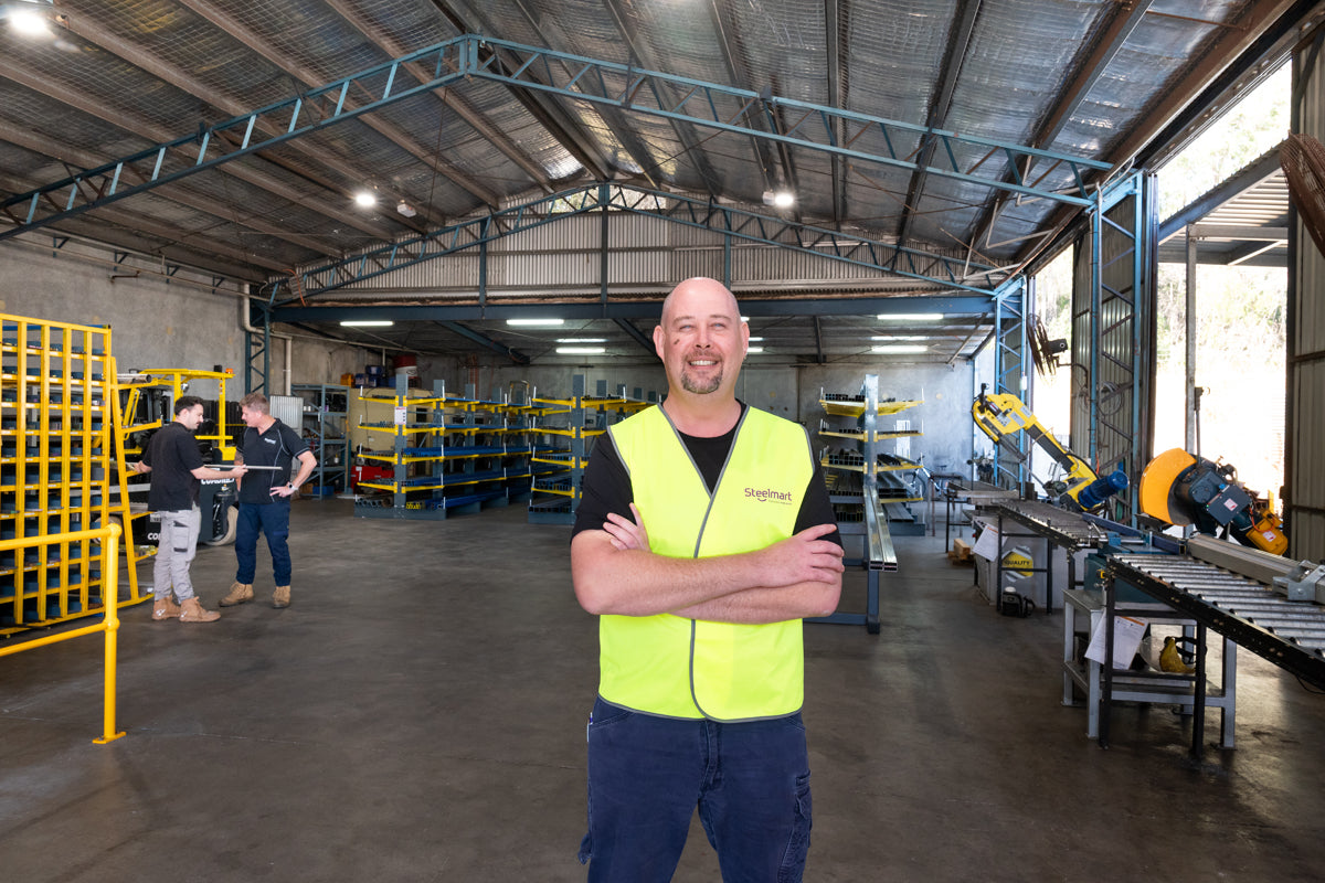 steel_warehouse_customers_choosing_steel_sales_person_smiling_foreground_wearing_yellow_hi_vis_safety_vest